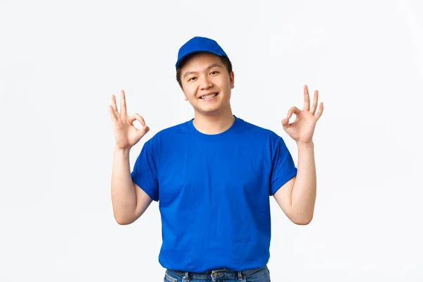 Sonriente mensajero asiático seguro de sí mismo en uniforme azul, guiño y mostrando gesto bien. El tipo de la entrega dice OK, garantiza la seguridad y el envío rápido de paquetes, el hombre que trabaja en el correo asegura al cliente — Foto de Stock