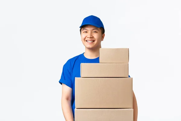 Compras online, concepto de envío rápido. Agradable mensajero asiático sonriente, repartidor en uniforme azul llevar paquetes a la puerta, llevar cajas con órdenes y de pie fondo blanco —  Fotos de Stock