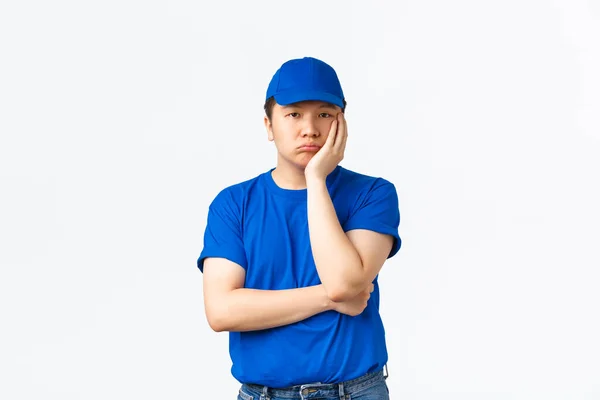 Πορτρέτο του βαριέται και χωρίς διασκέδαση ασιατική courier σε μπλε στολή, καπέλο και t-shirt, άπαχο πρόσωπο στο χέρι και αναζητούν αδιάφοροι. Παράδοση άνθρωπος πολύ δυσαρεστημένος με κάτι, στέκεται λευκό φόντο — Φωτογραφία Αρχείου