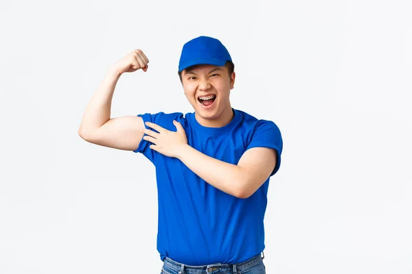 Sassy mosolygós ázsiai futár kék egyenruhában, rugalmas bicepsszel magabiztos. A futár önbizalmát fejezi ki, megmutatva, milyen erős a karja, nehéz ügyfelek csomagok, álló fehér háttér — Stock Fotó
