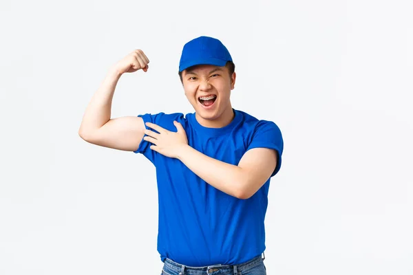 Corredor asiático sonriente descarado en uniforme azul, bíceps flexibles confiados. El repartidor expresa confianza, mostrando lo fuertes que son sus brazos, lleva paquetes de clientes pesados, de pie fondo blanco — Foto de Stock