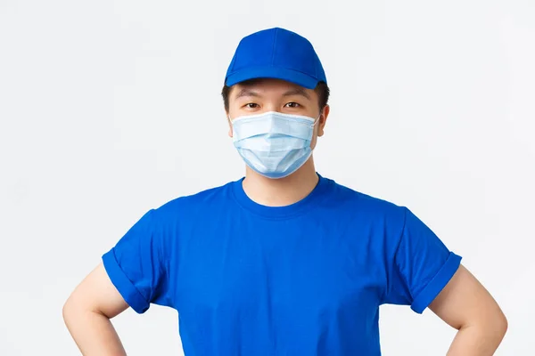 Onlineshopping, leverans och coronavirus pandemi, förhindra virus koncept. Närbild av säker leende asiatisk manlig kurir i blå uniform, medicinsk mask, ger säker pakethantering — Stockfoto