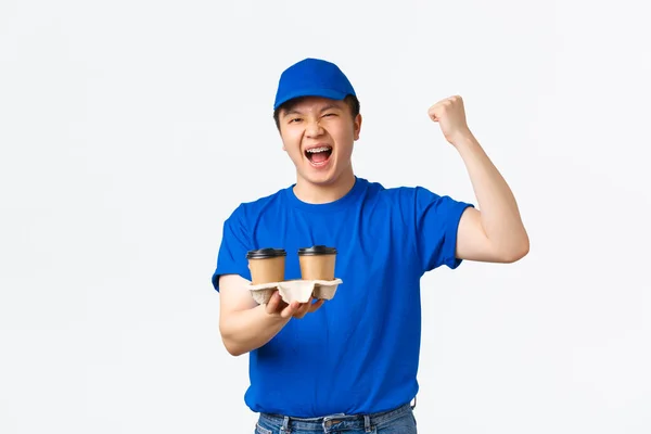 Online orders, takeaway and employees concept. Triumphing cheerful asian male courier in blue uniform bring coffee to client home without spill, rejoicing, shouting hooray or yes, white background — Stock Photo, Image