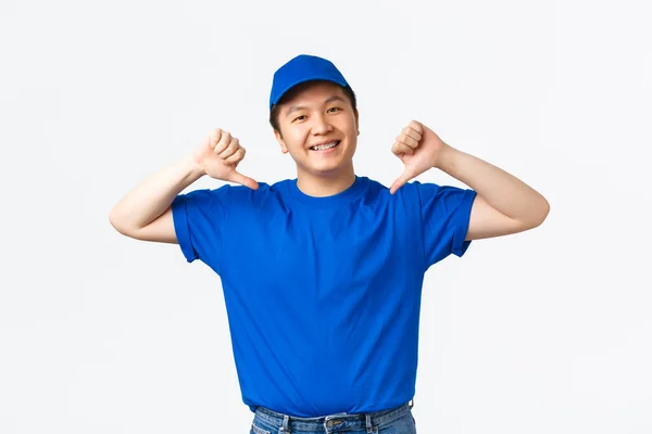 De aspecto amistoso sonriente y confiado asiático repartidor en uniforme azul señalándose a sí mismo. Mensajero promover su servicio de empresa, garantía de envío rápido y seguro, de pie fondo blanco —  Fotos de Stock