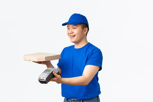 Conceito de compras, pagamento e entrega sem contato. Sorrindo amigável-olhando asiático courier em azul uniforme, entregando caixa de pizza, ordem para o cliente e dar terminal POS, fundo branco — Fotografia de Stock