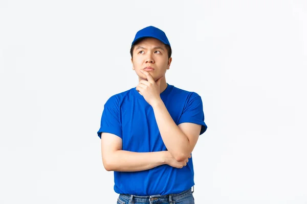 Στοχαστικός Ασιάτης ντελιβεράς με μπλε μπλουζάκι και καπέλο που κάνει επιλογές, σκέφτεται ή συλλογίζεται το σχέδιο, κοιτάζει την πάνω αριστερή γωνία σοβαρά, στέκεται λευκό φόντο, αποφασίζει τι θα κάνει — Φωτογραφία Αρχείου