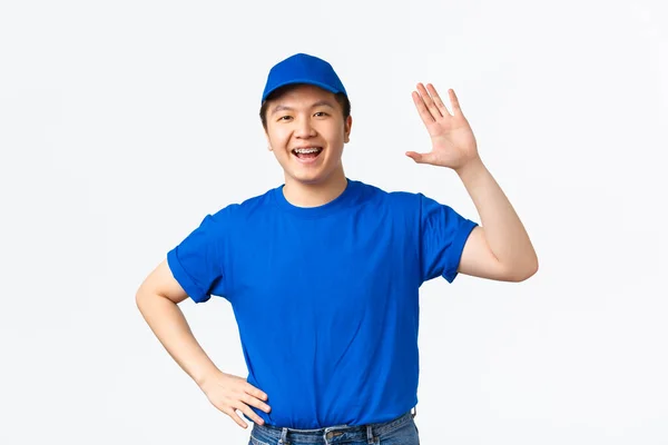 Φιλικός Ασιάτης κούριερ με μπλε στολή χαμογελά και παραιτείται από το χέρι για να πει γεια, χαιρετώντας πελάτη φέρει την αγορά στο κατώφλι του πελάτη. Χαρούμενος ντελιβεράς κάνει γεια χειρονομία, λευκό φόντο — Φωτογραφία Αρχείου