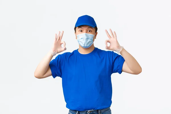 Απρόσκοπτη παράδοση, μεταφορά covid-19, πρόληψη της έννοιας του ιού. Φιλικός Ασιάτης κούριερ με μπλε στολή, ιατρική μάσκα και γάντια εξασφαλίζουν την ποιότητα και την ασφάλεια της αποστολής, δείχνοντας εντάξει χειρονομία — Φωτογραφία Αρχείου