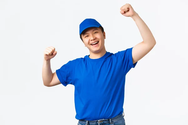 Portret van de succesvolle winnende Aziatische koerier in blauw uniform verheugen, breed glimlachen en dansen van triomf, vieren overwinning of prestatie, vuist pomp, zeggen hoera, witte achtergrond — Stockfoto