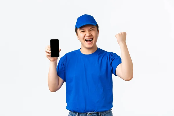 Technology, online shopping and shipping concept. Winning happy asian delivery man celebrating achievement, standing in courier uniform, showing fist pump and mobile phone screen, white background — Stock Photo, Image