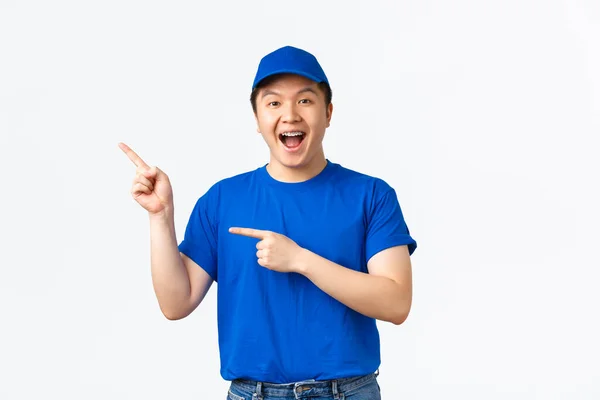 Wesoły młody azjatycki kurier w niebieskim mundurze wskazujący palcem górny lewy róg, reklama firmy przewozowej, przesyłka paczek. Słodki dostawca coś promować — Zdjęcie stockowe