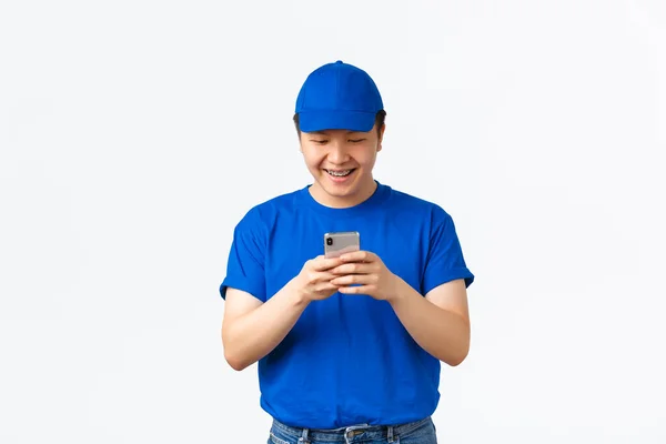 Tecnologia, compras on-line e conceito de transporte. Sorrindo feliz asiático entregador homem com aparelho, vestindo azul courier uniforme, olhando para tela do smartphone, conversando ou usando aplicativo — Fotografia de Stock