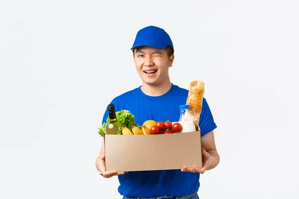 Online shopping, food delivery and shipment concept. Winking cute asian male courier in blue uniform provide fast shipment, bring groceries order to client house, handing box and smiling — Stock Photo, Image