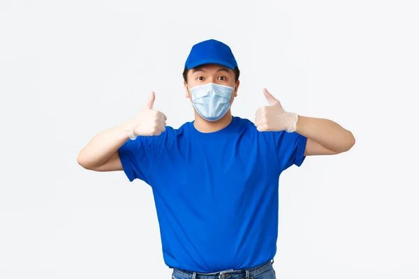 Contactless delivery, covid-19 shipping, preventing virus concept. Asian male courier in blue uniform, wear medical mask and gloves, show thumbs-up in approval, guarantee fast and safe shipment — Stock Photo, Image