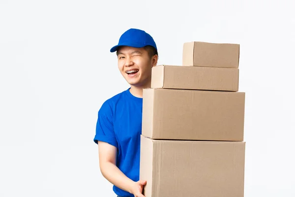 Compras online, conceito de transporte rápido. Cheeky sorrindo asiático courier em azul uniforme segurando caixas com ordens do cliente, piscar amigável no cliente, fornecer entrega rápida, de pé fundo branco — Fotografia de Stock