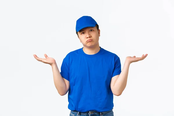 Porträtt av ryckiga och aningslösa asiatiska leverans man i blå uniform höja händerna i sidled omedvetna. Courier vet ingenting, står ledsen och dyster utan svar, vit bakgrund — Stockfoto