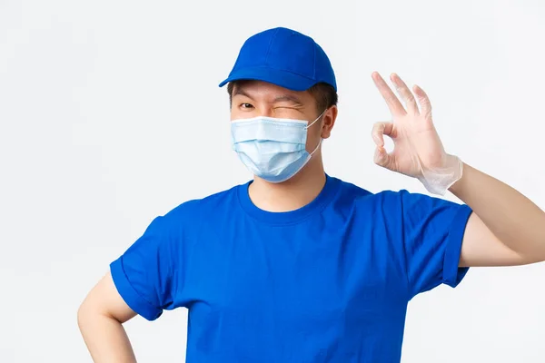 Online αγορές, παράδοση και coronavirus πανδημία, την πρόληψη της έννοιας του ιού. Ασιάτης κούριερ με μπλε στολή, ιατρική μάσκα και γάντια, παρέχει ασφαλή παράδοση κατά τη διάρκεια covid-19, δείχνουν εντάξει χειρονομία — Φωτογραφία Αρχείου