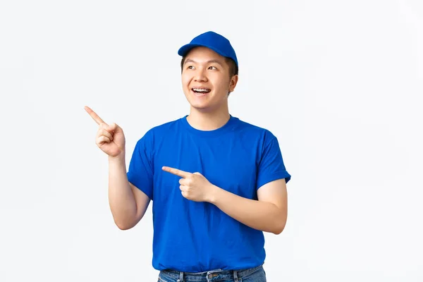 Drömmande glad leende kurir med hängslen, bär blå uniform, ser nöjd och fascinerad, pekar finger övre vänstra hörnet på reklam, stående vit bakgrund — Stockfoto