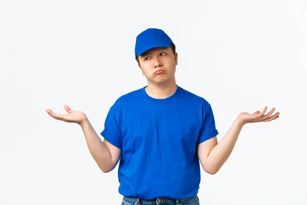Een besluiteloze Aziatische jonge bezorger in blauw uniform, die zich schuil houdt en verbaasd wegkijkt, handen zijwaarts spreidt zonder het te weten. Koerier staande verbijsterd over witte achtergrond — Stockfoto