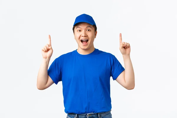 Opgewonden lachende Aziatische jongeman werkt als koerier, draagt blauwe uniform pet en t-shirt, wijst vingers omhoog en ziet er vrolijk uit, maakt aankondiging, promotie aanbod, witte achtergrond — Stockfoto