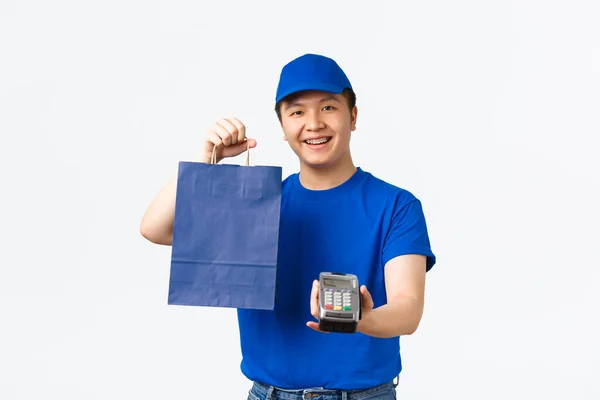 무심 한 지불, 쇼핑, 가정 배달 개념. 파란 유니폼을 입은 우호적으로 보이는 아시아인 택배, 소포, 고객을 위해 패키지 , POS 터미널을 넘겨 — 스톡 사진