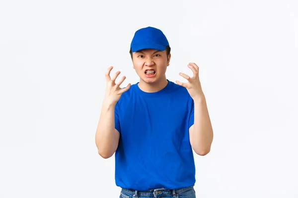 Pissed-off and angry asian delivery man in blue uniform shaking hands near head, grimacing mad and outraged. Courier looking distressed, tired of rude customers, standing white background — Stockfoto