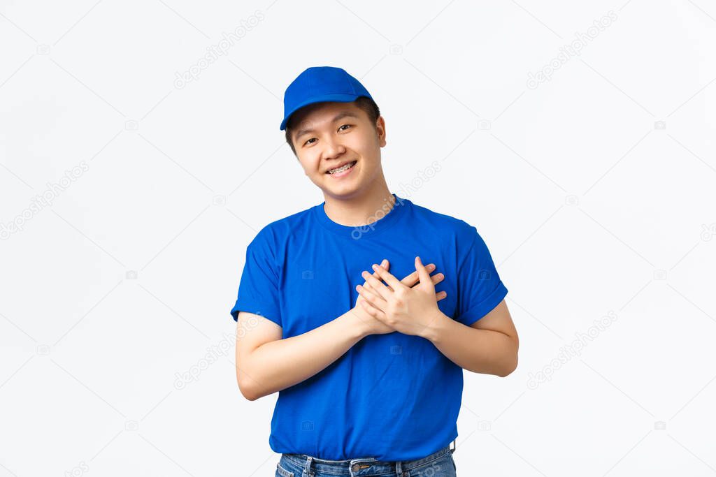 Touched cute asian courier in blue uniform love his clients, care for customer parcels, holding hands on heart and smilig flattered. Delivery guy appreciate praises of his work, white background