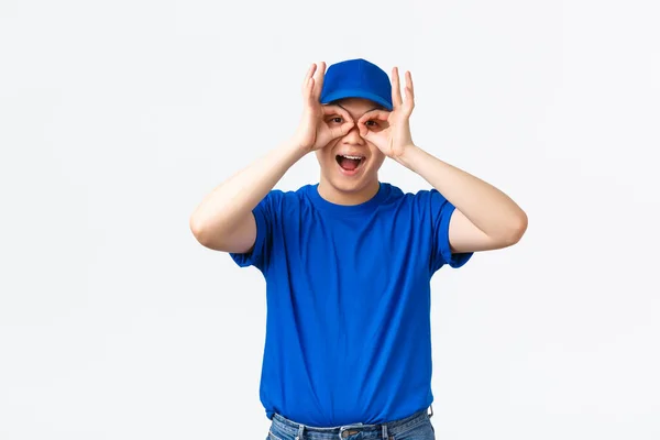 Vrolijke grappige Aziatische bezorger in blauw uniform pet en t-shirt, het maken van goede gebaren, handbril over de ogen en glimlachende upbeat. Koerier rommelen rond staan speels over witte achtergrond — Stockfoto