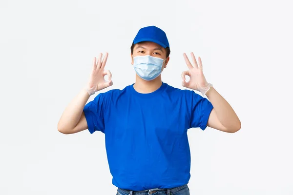 Απρόσκοπτη παράδοση, μεταφορά covid-19, πρόληψη της έννοιας του ιού. Φιλικός Ασιάτης κούριερ με μπλε στολή, ιατρική μάσκα και γάντια εξασφαλίζουν την ποιότητα και την ασφάλεια της αποστολής, δείχνοντας εντάξει χειρονομία — Φωτογραφία Αρχείου