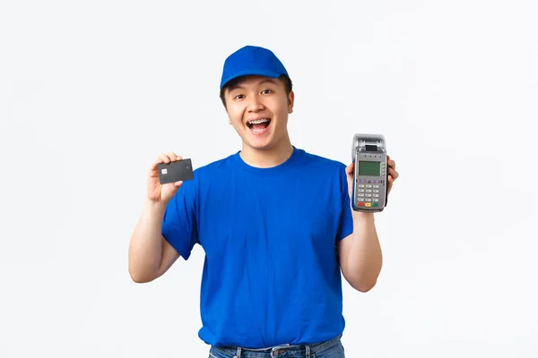 Compras sem contato, pagamento e conceito de funcionários. Sorrindo, mensageiro de aparência amigável, homem de entrega em uniforme azul mostrando cartão de crédito e terminal de POS, de pé fundo branco — Fotografia de Stock