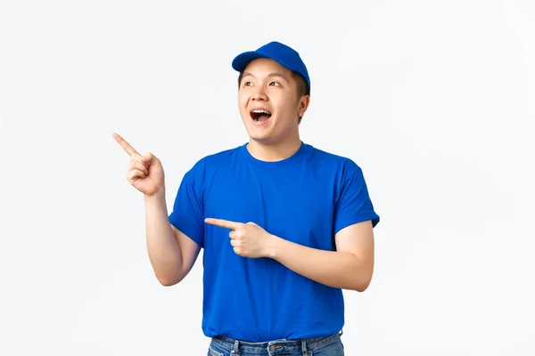 Spännande och förvånad asiatisk leverans kille i blå t-shirt och mössa, företagets uniform, pekar och ser övre vänstra hörnet med euforisk, glada uttryck, se awesome promo, vit bakgrund — Stockfoto