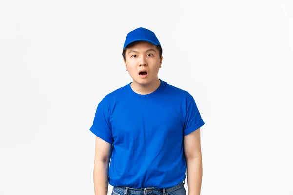 Πορτρέτο του σοκαρισμένος και έκπληκτος ασιατικό delivery τύπος σε μπλε καπέλο στολή και t-shirt αντιδρούν σε κάτι που κόβει την ανάσα, πτώση σαγόνι και άφωνος κάμερα πάνω από το λευκό φόντο — Φωτογραφία Αρχείου