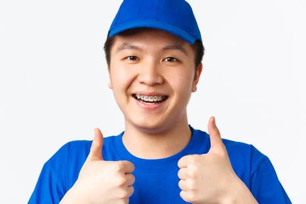 Online winkelen, snelle verzending, medewerkers en home delivery concept. Close-up van vrolijke glimlachende Aziatische mannelijke koerier met beugel, in blauw uniform, met duimen-up in goedkeuring, aanbevelen — Stockfoto