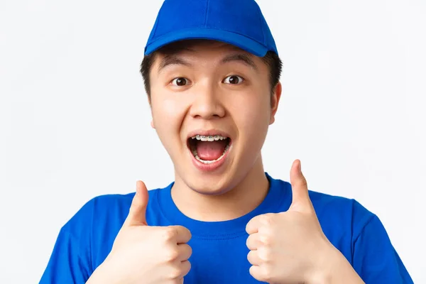 オンラインショッピング、高速配送、従業員と家庭配達の概念。興奮した幸せなアジアの男性の宅配便のクローズアップ青い制服,親指アップと面白い見て,白い背景 — ストック写真