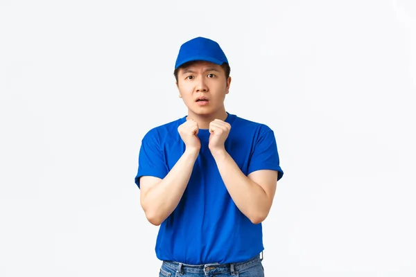 Worried and concerned asian delivery guy in blue uniform looking indecisive and scared, holding hands pressed to chest and making nervous expression, standing white background