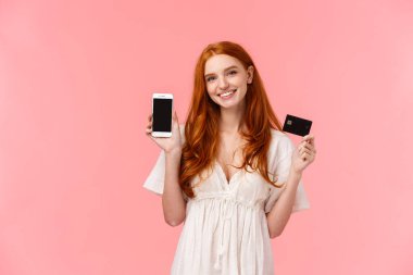 Mobil ekranda uygulamayı tanıtan kız. Neşeli, güzel kızıl saçlı beyaz kadın kredi kartı tutuyor ve akıllı telefon ekranını gülümseyerek gösteriyor. Uygulama ya da internet mağazası tanıtıyor.