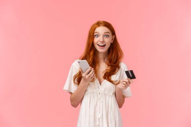 Etkilenmiş ve heyecanlı, beyaz elbiseli mutlu kızıl saçlı kız, ne kadar kolay satın aldığına şaşırmış, elinde akıllı telefon ve kredi kartı, gülümsüyor ve merak uyandıran kamera, pembe arka plan.