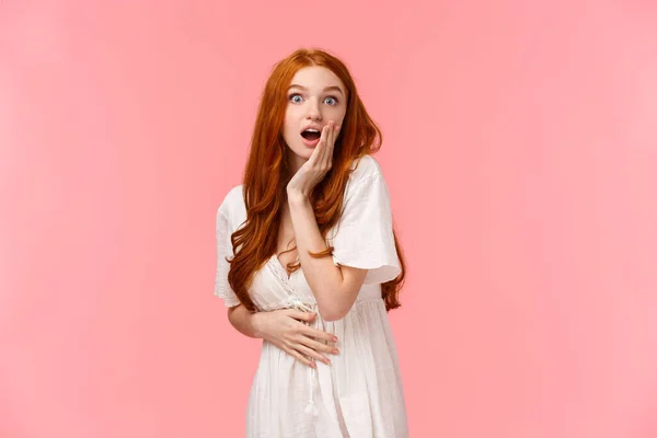 Waist-up portrait surprised and curious, astonished redhead girl heard amazing news, gossiping with girlfriend on party, open mouth impressed, touch cheek look wondered, pink background — 스톡 사진
