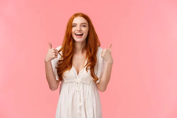 Cintura-up retrato alegre, mulher ruiva otimista em vestido branco, mostrando polegares para cima e sorrindo na aprovação, como produto, recomendo, expressar opinião positiva, de pé fundo rosa — Fotografia de Stock