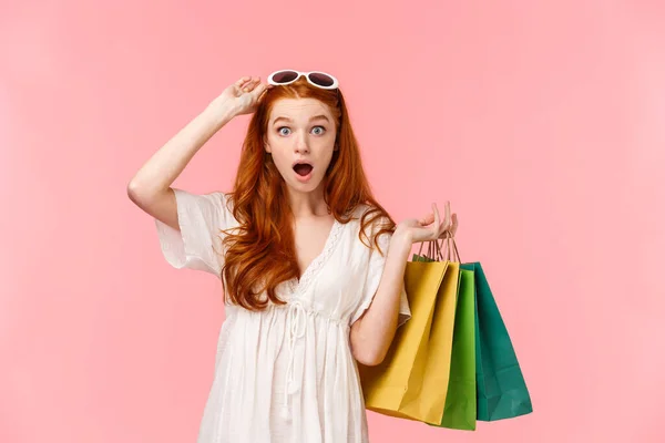 Waist-up portrait impressed, excited and surprised ginger girl, foxy curly hair, take-off sunglasses, seeing amazing discounts in store, hurry up to buy something, holding bags with shopping goods