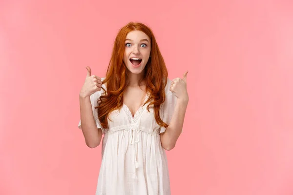 Réductions, achats et concept de personnes. Femme rousse gaie étonnée de l'événement merveilleux, assister à un concert impressionnant, montrer pouces levés et souriant étonné, fond rose — Photo