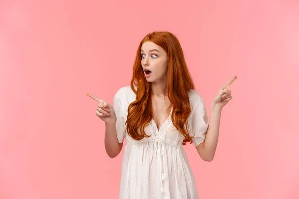 Amazed, curious and excited redhead female in white dress, seeing something interesting, checking out great offer, awesome opportunity, pointing fingers left and right, two choices — 스톡 사진