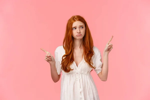 Waist-up portrait innocent, cute and timid redhead indecisive girl, dont know what do, pouting feeling sad, gloomy look around, pointing fingers left and right, pink background — 스톡 사진