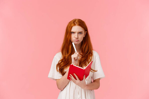 Serious-looking grumpy redhead woman frowning as thinking, pouting troubled, solving serious riddle, holding red notebook and touching chin with pen, pondering over homework, pink background