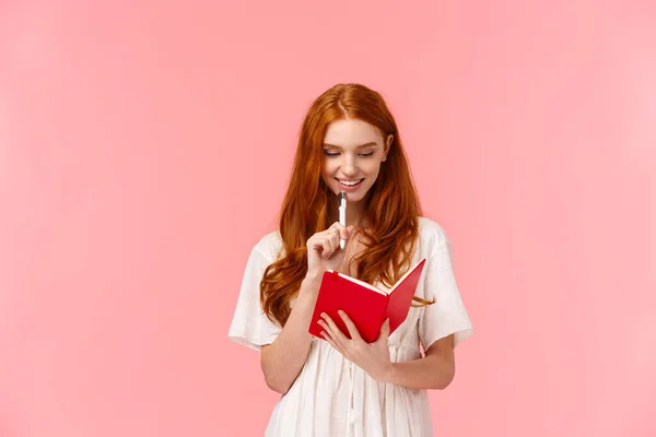 Nachdenklich und glücklich, lächelnd hübsche rothaarige Frau in weißem Kleid, beim Denken das Kinn mit Stift anfassen, Einkaufen oder To-do-Liste machen, etwas in süßes rotes Notizbuch schreiben, rosa Hintergrund — Stockfoto