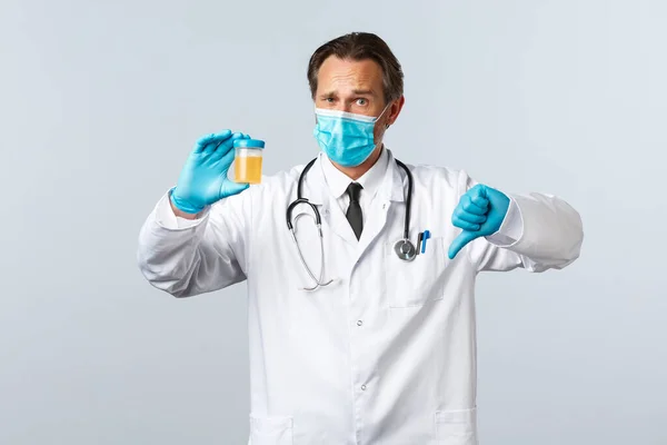 Covid-19: 바이러스 예방, 의료 종사자 및 예방 개념. 의료용 마스크와 장갑을 끼고 있는 정신과 의사는 소변 샘플을 보여 주고, 엄지손가락을 내리는 것은 안 좋은 검사 결과를 보여 주고, 하얀 배경을 보여 줍니다. — 스톡 사진