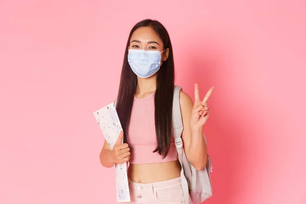 Safe tourism, travelling during coronavirus pandemic and preventing virus concept. Happy asian girl travel in pandemic covid-19 with medical mask, holding map, show peace sign, tourist going abroad