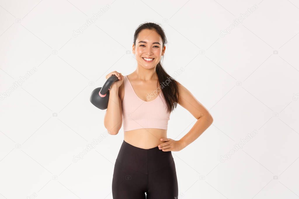 Sport, wellbeing and active lifestyle concept. Smiling strong and slim asian fitness girl, female athlete holding kettlebell and looking carefree, gaining muscles at gym, standing white background