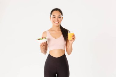Spor, refah ve aktif yaşam tarzı konsepti. Portakal suyu ve salata tutan sağlıklı ve formda Asyalı bir sporcunun portresi. Sabah kahvaltısı antrenmandan önce, beyaz arka plan.