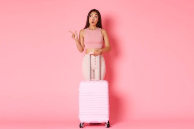 Seyahat, tatil ve tatil konsepti. Etkilenmiş Asyalı turist kız çocuğu. Yurt dışına seyahat ederken etrafı gezerken hayret içinde bakıyor. Pembe arka planda bavulla yürüyor.